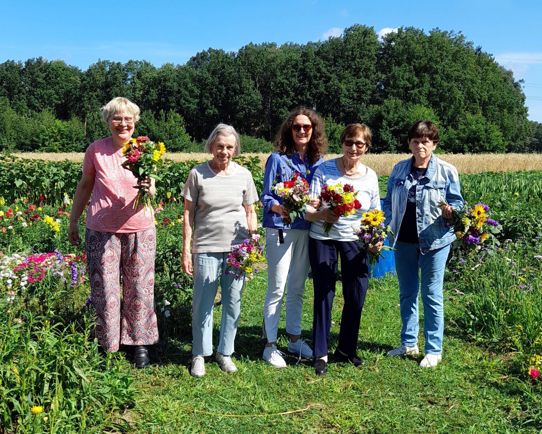 Fünf Frauen stehen bei strahlendem Sonnenschein auf einer Blumenwiese und halten frisch gepflückte Blumensträuße in den Händen. Im Hintergrund sind Felder und ein Waldrand zu sehen – die Szene vermittelt Naturverbundenheit, Freude und Gemeinschaft.